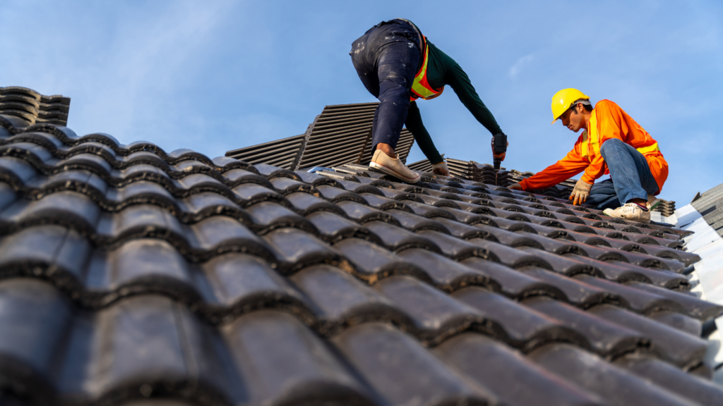 Photo of two roofers in safety gear installing or repairing a roof with dark shingles under a clear blue sky.