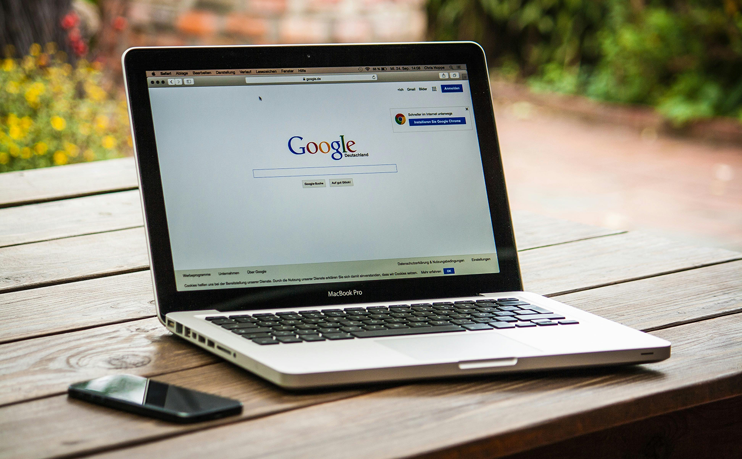 An open MacBook Pro displaying the Google search homepage sits on a wooden table next to a black smartphone, with a blurred garden in the background.